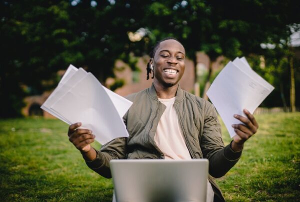Black man in park on laptop smiling holding papers.