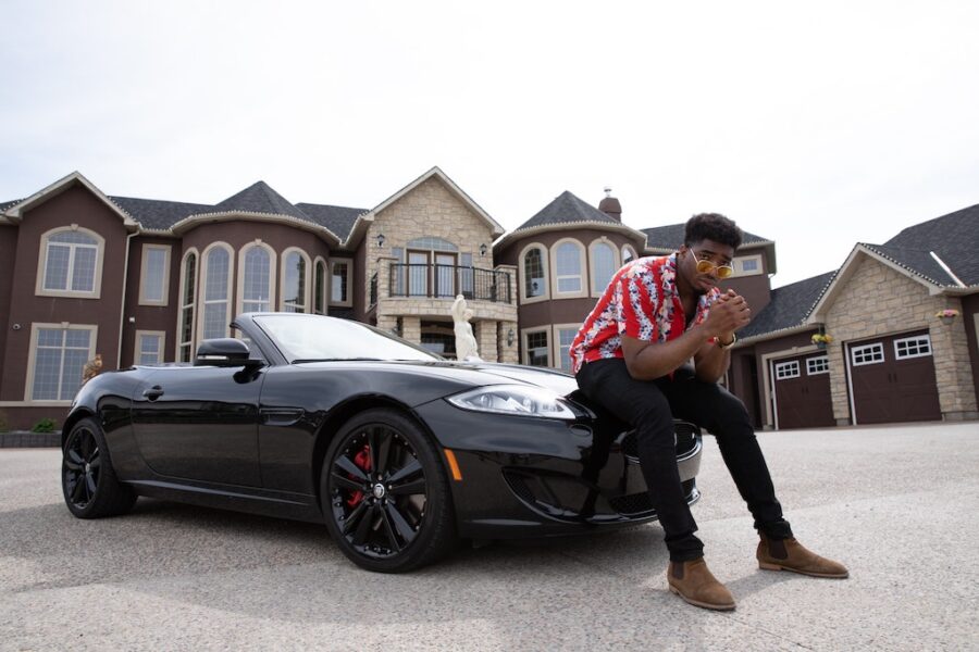 Black millionaire sitting on convertible car hood in front of a mansion.