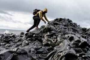 a black man in a yellow coat climbing on black rocks near the ocean