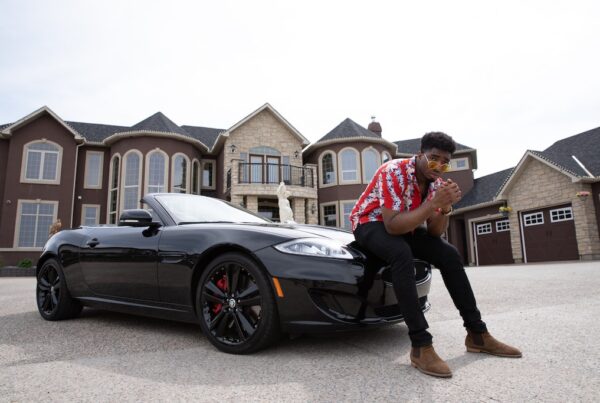 Black millionaire sitting on convertible car hood in front of a mansion.